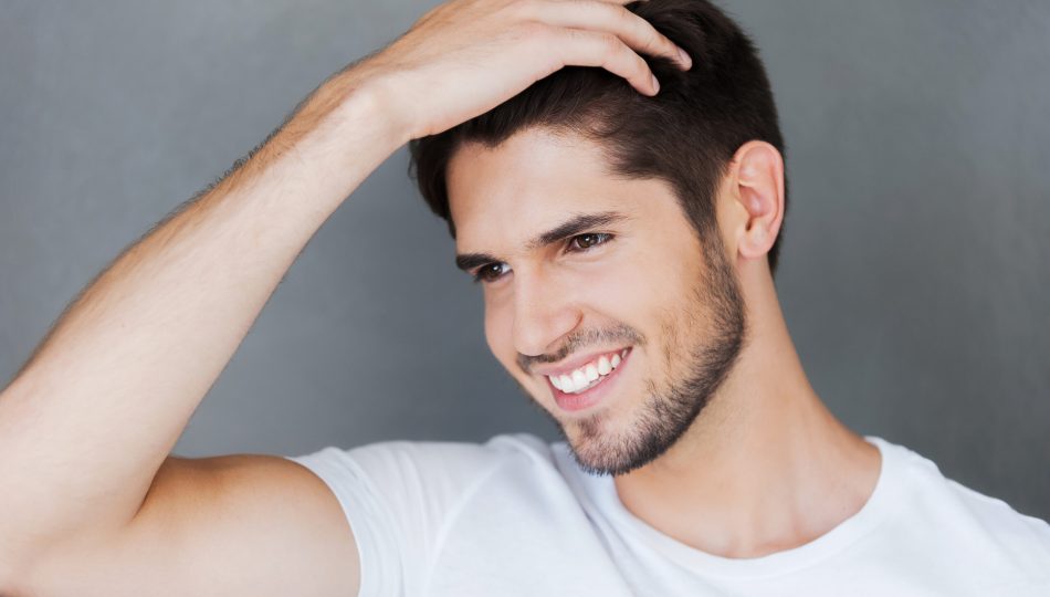 Carefree handsome. Happy young man holding hand in hair and looking away while standing against grey background